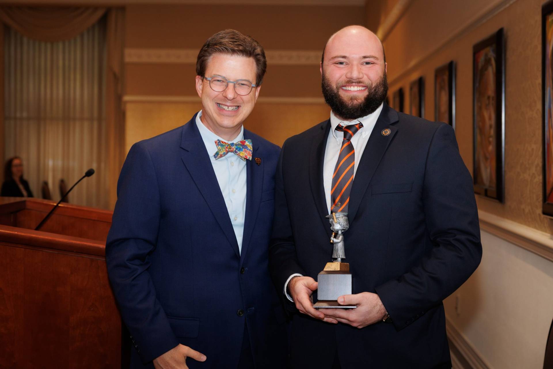 Two men in suits stand indoors; the man on the right holds a trophy and both are smiling at the camera.