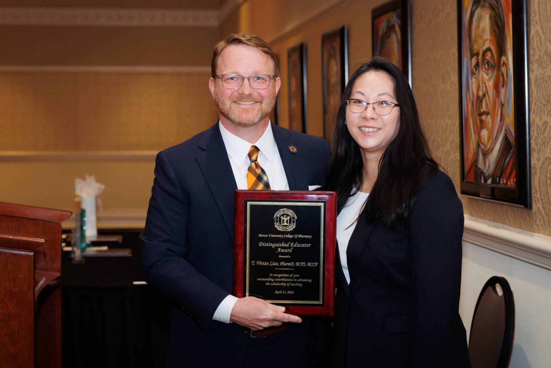 Two people in business attire stand indoors, one holding a plaque with an award inscription, smiling at the camera.