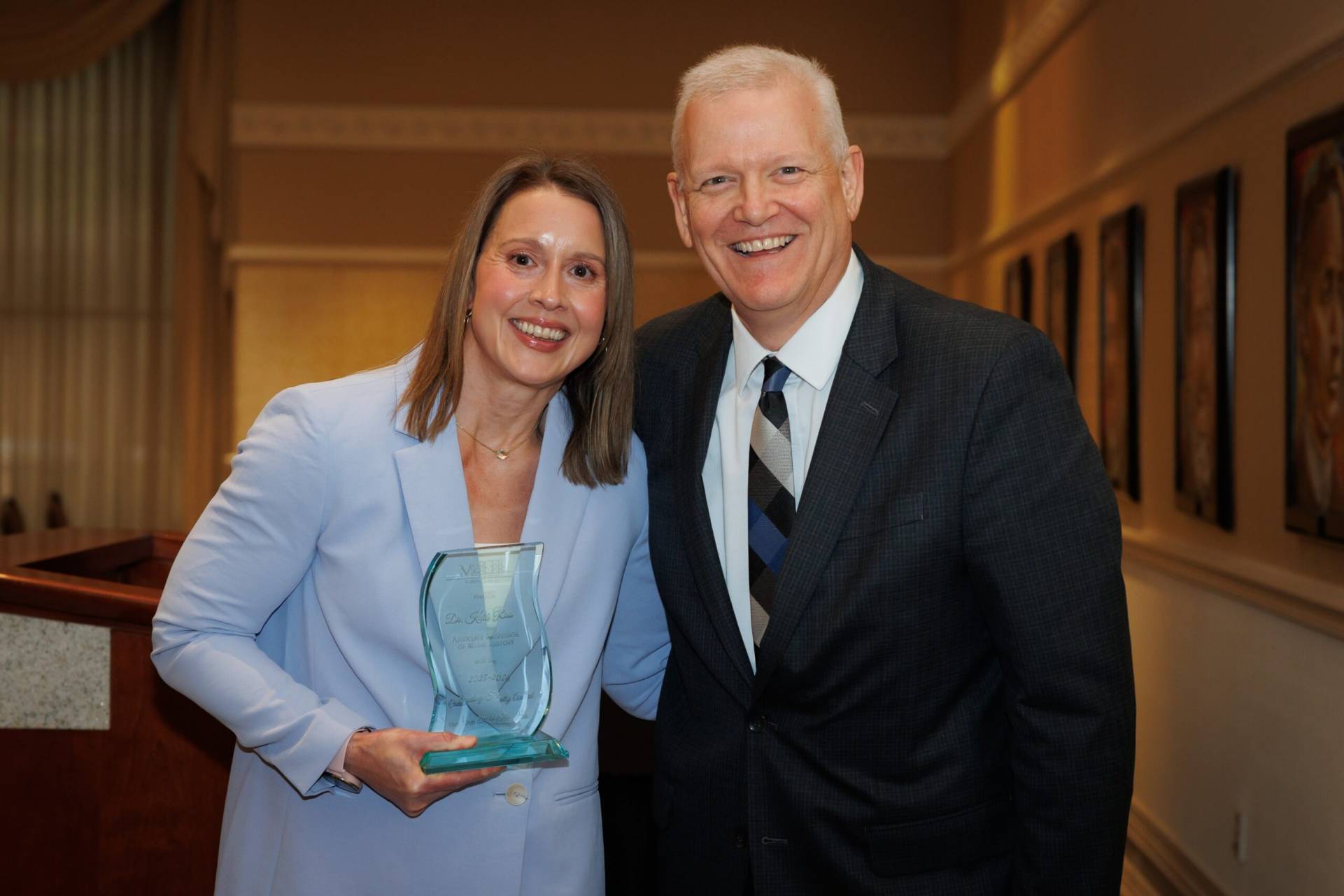 Two people stand indoors smiling; the woman on the left holds a glass award.