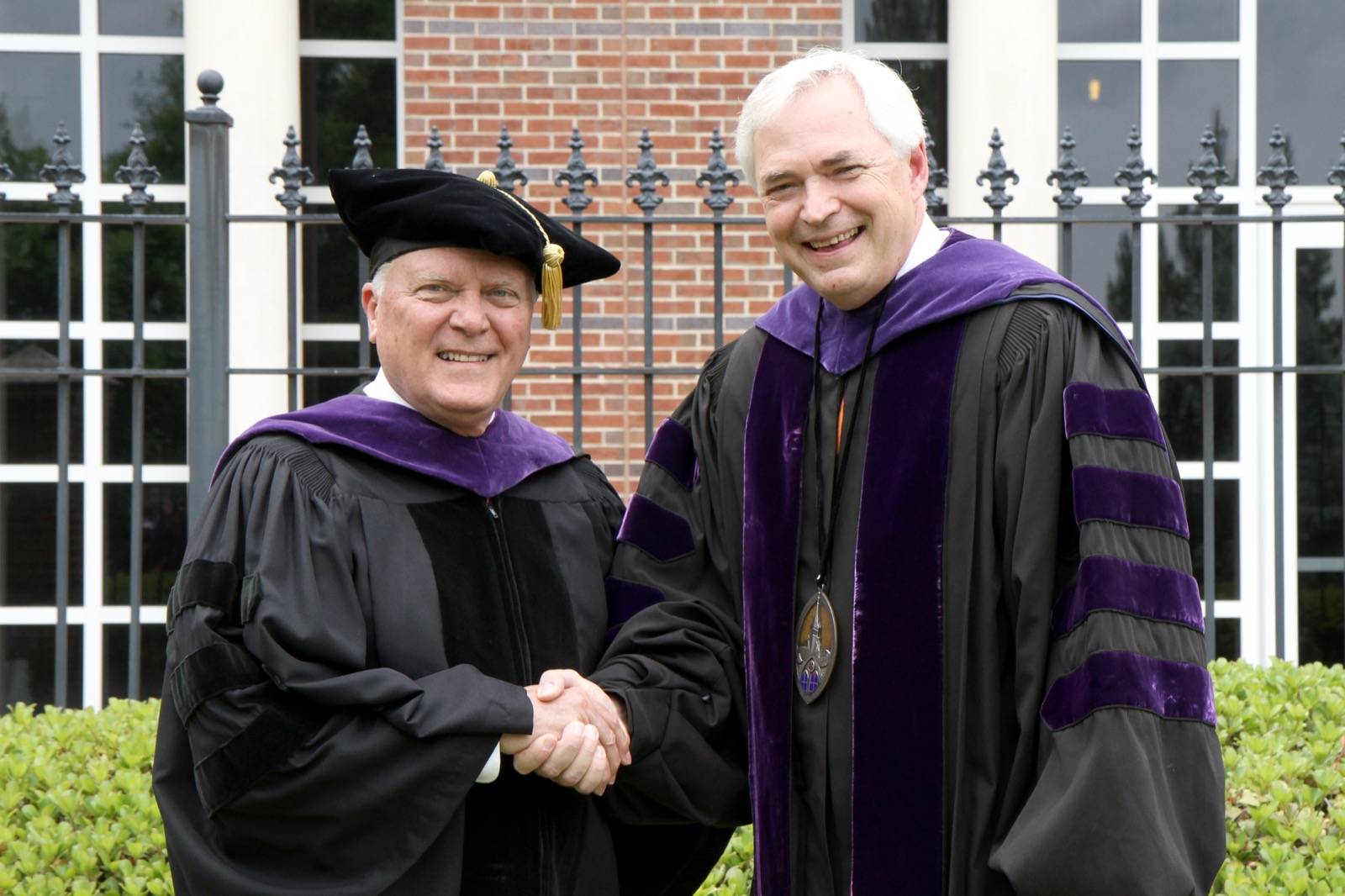 Two men in academic regalia shake hands and smile outside a building with brick walls and large windows.