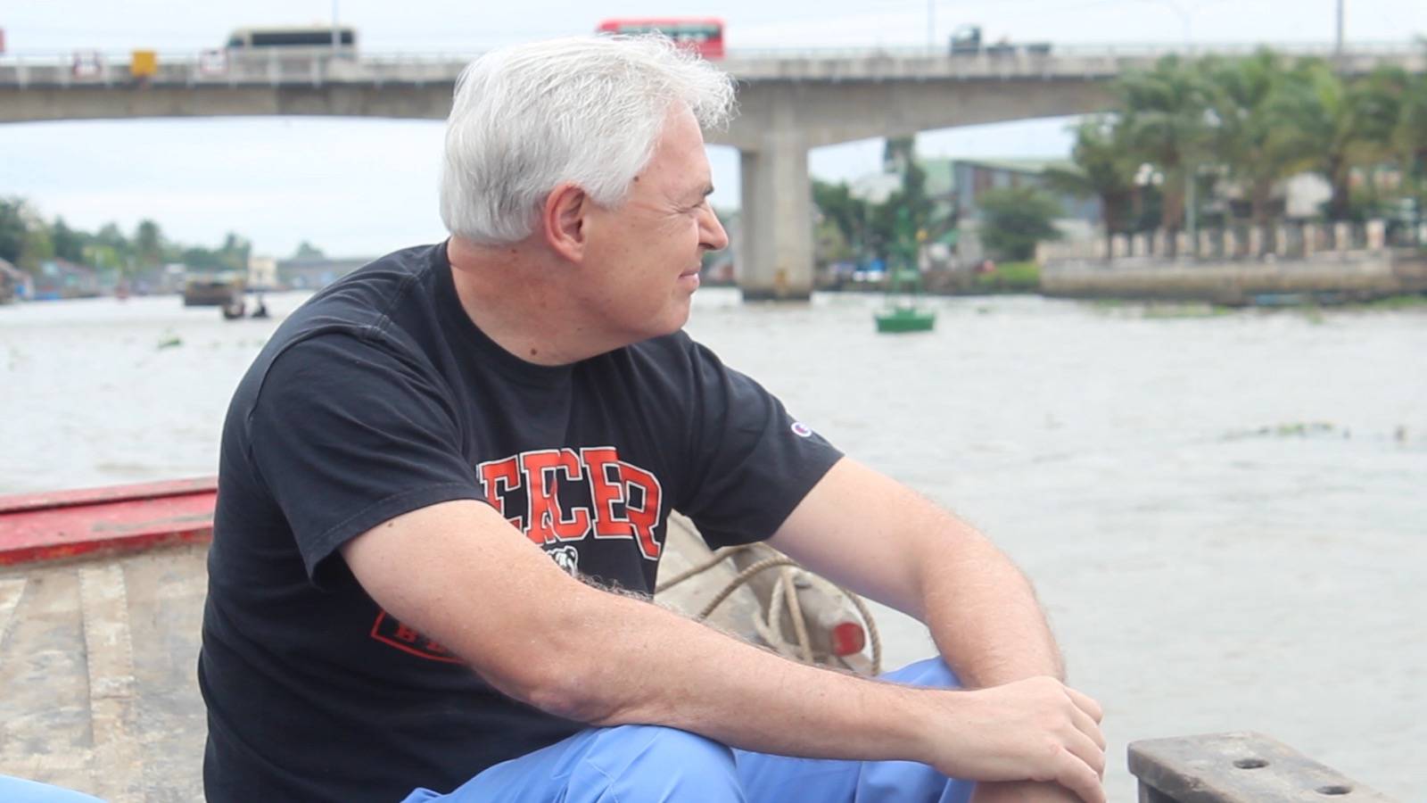 A man with gray hair sits on a boat, looking out over a river with a bridge and traffic in the background.