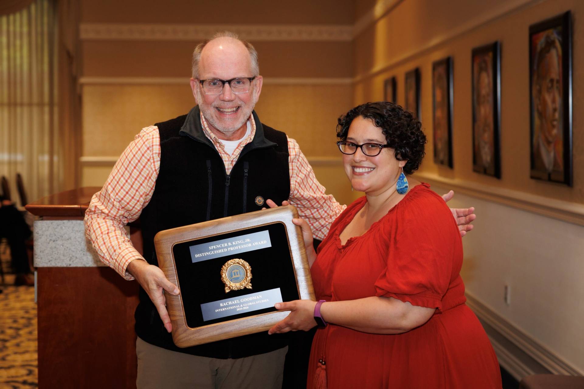Two people smiling as one presents the other with a framed distinguished service award in a gallery setting.