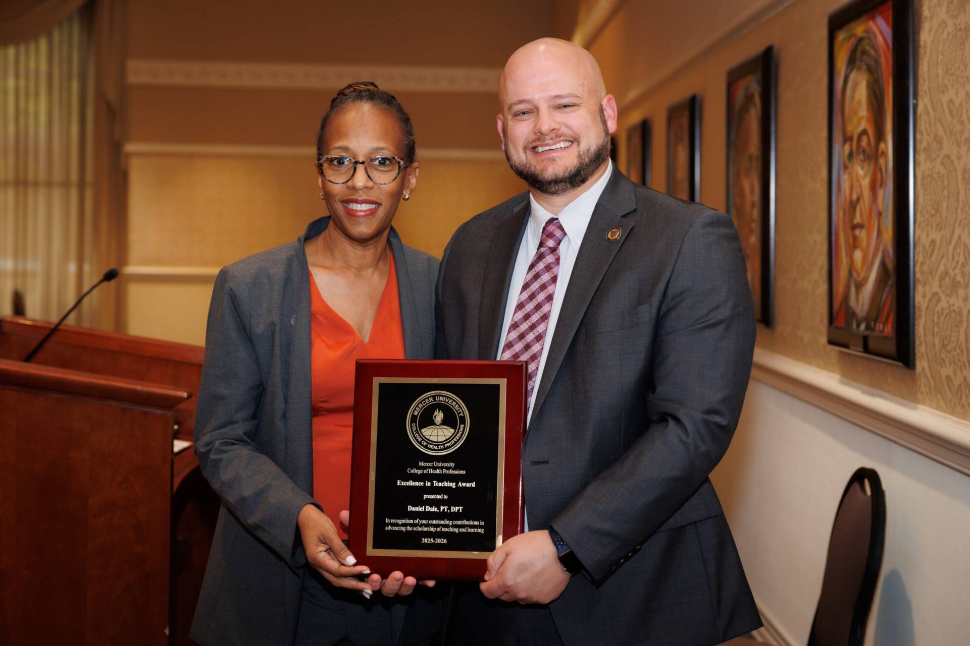 Two people in business attire hold a plaque reading Excellence in Teaching Award in an indoor setting.